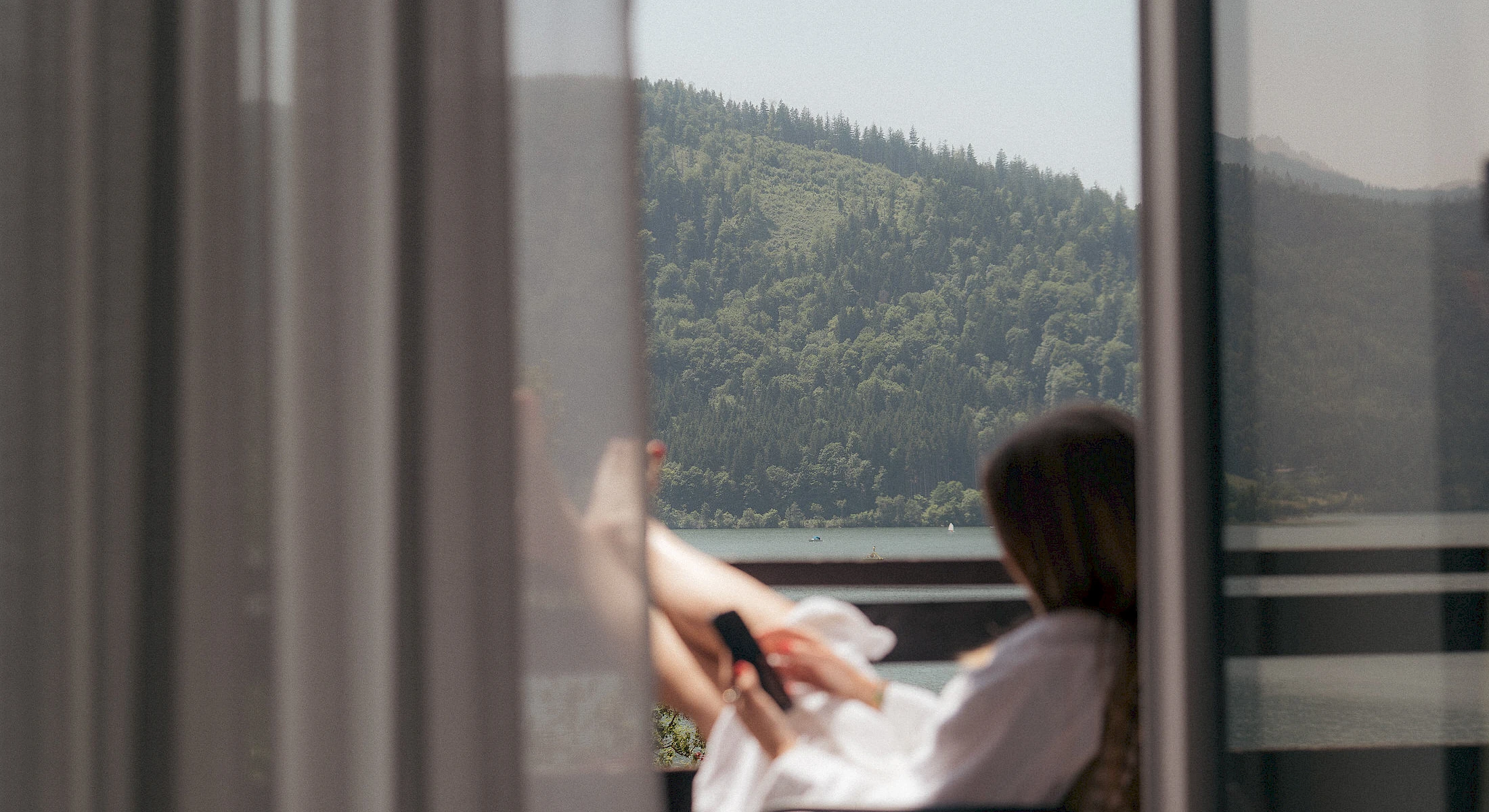 Frau im Bademantel sitzt am Balkon, trinkt Kaffe und genießt den Blick auf den Schliersee und die Berge.